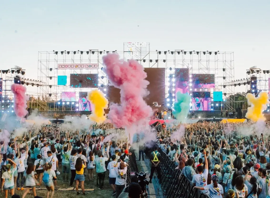 Público de un festival de música al aire libre frente al escenario, con nubes de polvo de colores